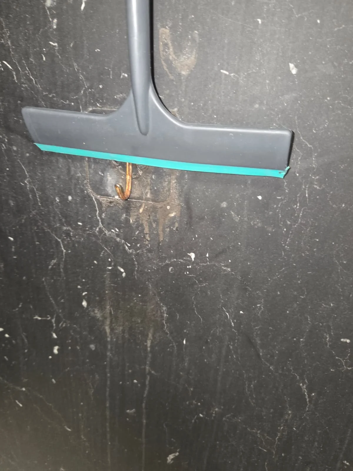 Close-up of a black cleaning squeegee with a blue edge, hanging on a rusty hook attached to a black, scratched surface.