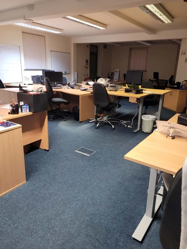 Empty office with multiple desks, office chairs, and desktop computers, illuminated by fluorescent ceiling lights.