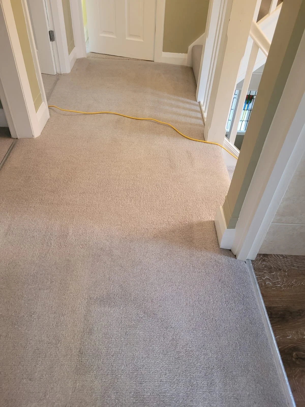 A hallway with beige carpet, yellow extension cord stretched across the floor, white door at the end, and stairway visible to the right.
