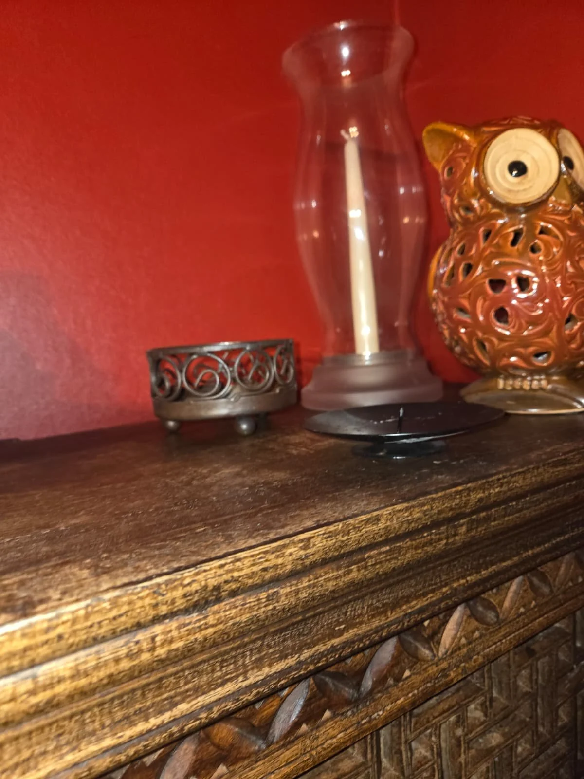 Decorative items on a wooden surface, including a glass candle holder with a candle, a small metal jewelry or trinket holder, and a ceramic owl-shaped decorative object, against a red wall background.
