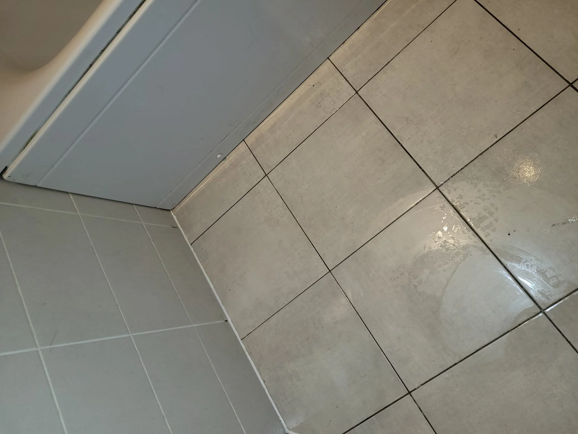 Bathroom floor with beige tiles and grout lines, next to a gray appliance, possibly a washing machine or dryer.
