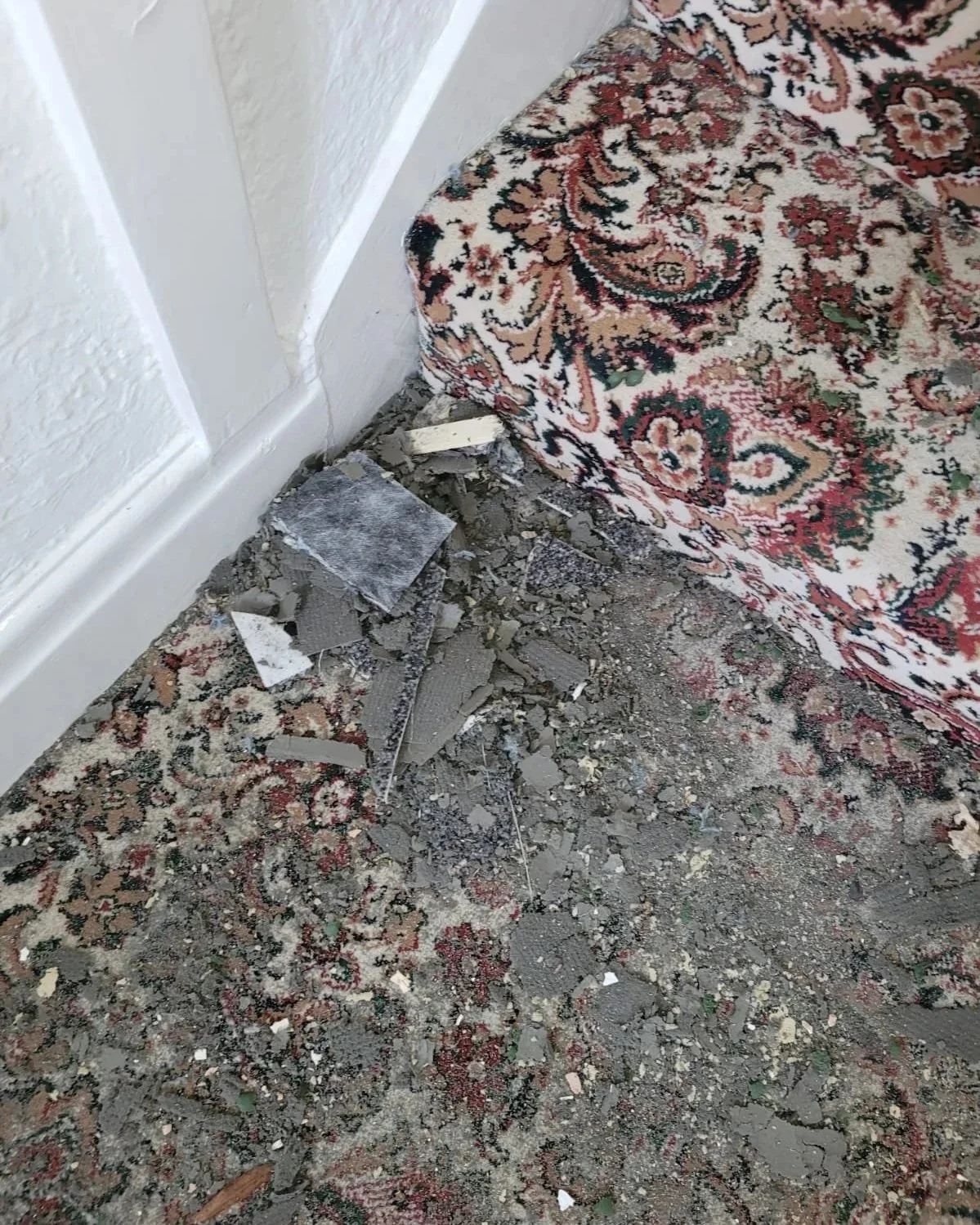 Broken pieces of drywall and debris on a dusty, patterned area rug next to a white wall and baseboard.
