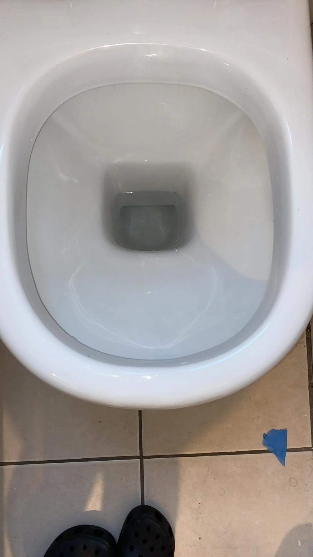 Looking down at a white toilet with the lid up, showing the inside of the bowl filled with water. The tiled floor and part of a person's shoes are visible at the bottom.