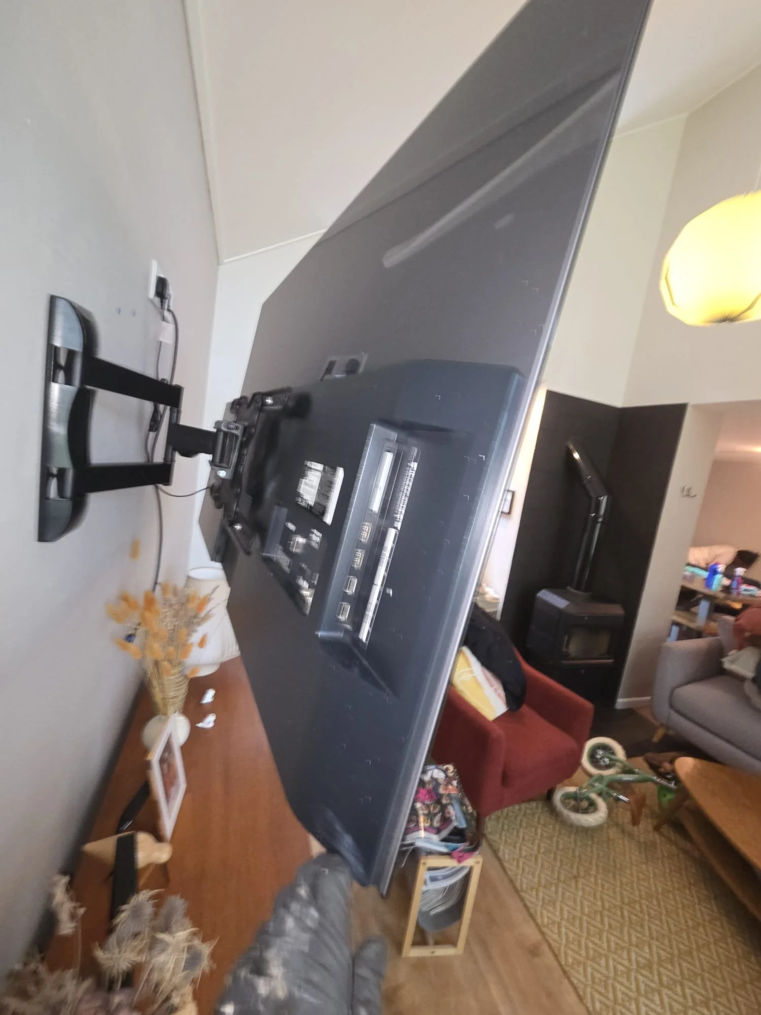 A large flat-screen TV mounted on a black stand in a living room. The TV is angled with a wall visible behind and a lamp in the upper right corner. The room has furniture including a red chair, a gray couch, a wood stove, and various decorative items