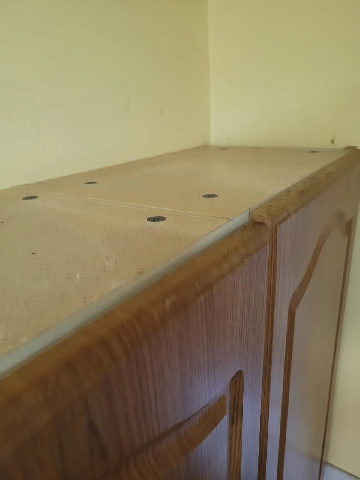 Close-up of a wooden cabinet with a light-colored, unfinished top surface against a yellow wall.