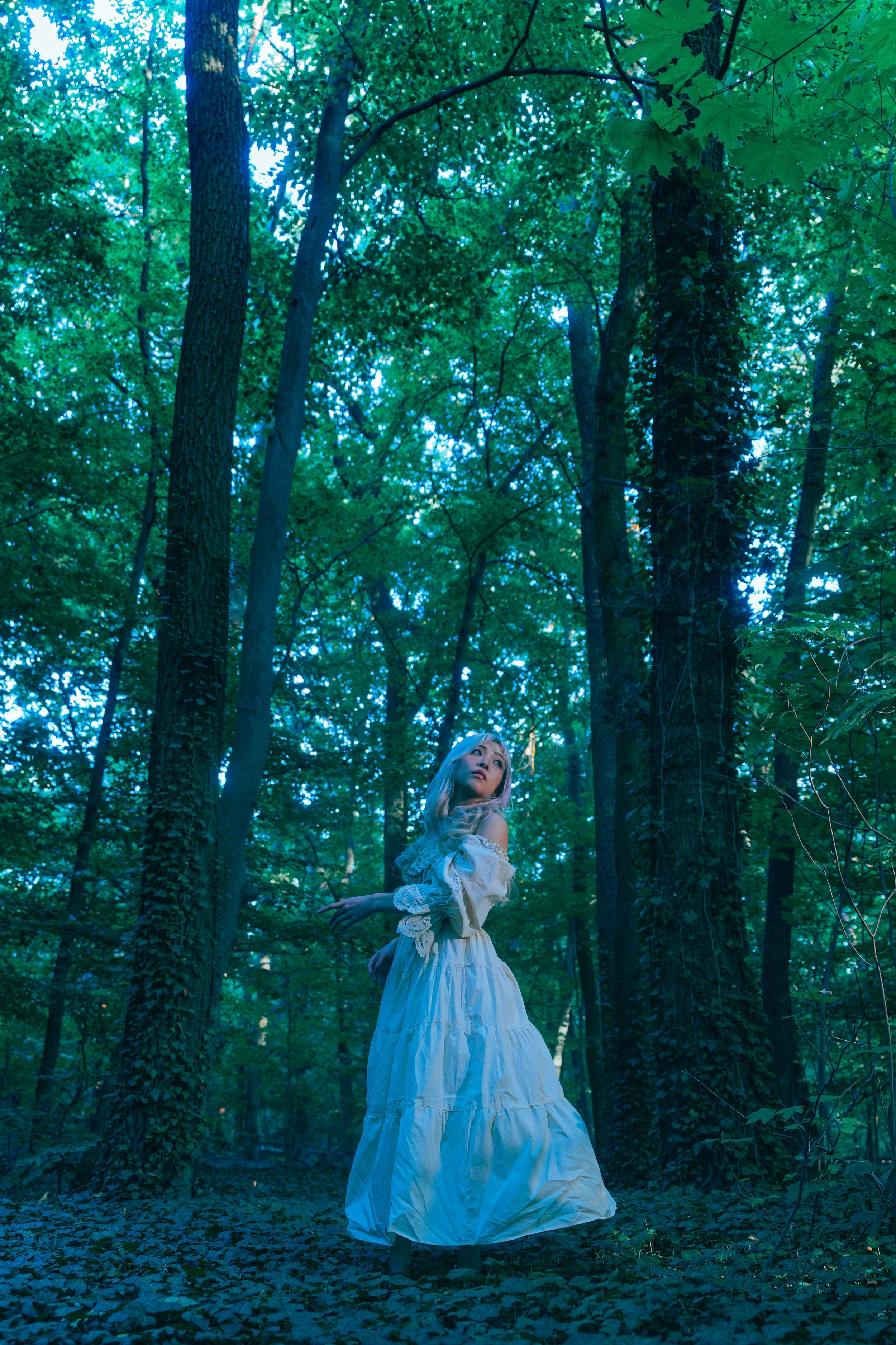 A woman in a flowing white dress stands in a dense green forest surrounded by tall trees and lush foliage. Cinematic portrait photography