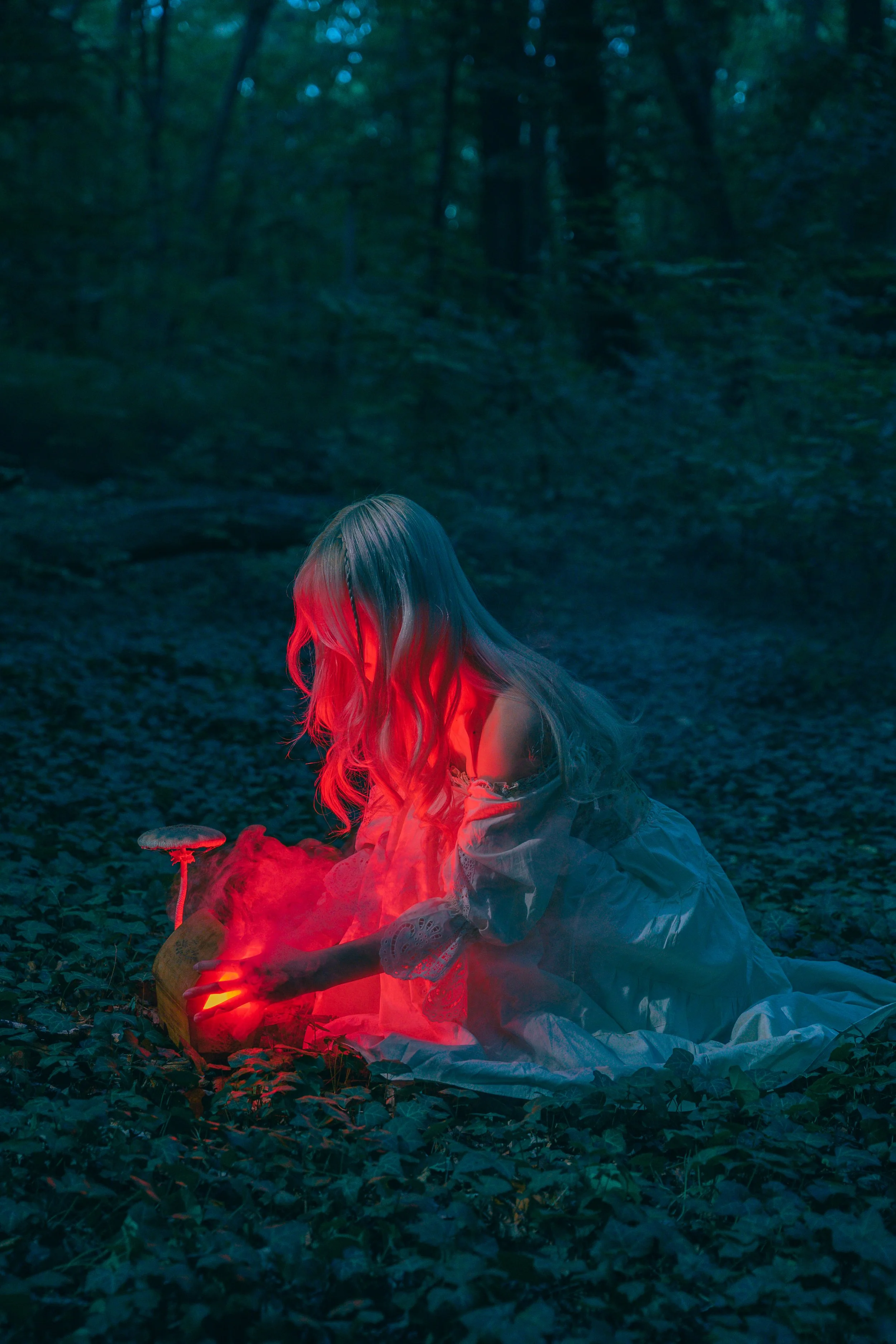 A woman with long, wavy blonde hair dressed in a vintage white dress is sitting on the forest floor with ivy, holding a glowing red lantern emitting smoke, surrounded by mushrooms, in a dark wooded area at dusk. Cinematic portrait photography