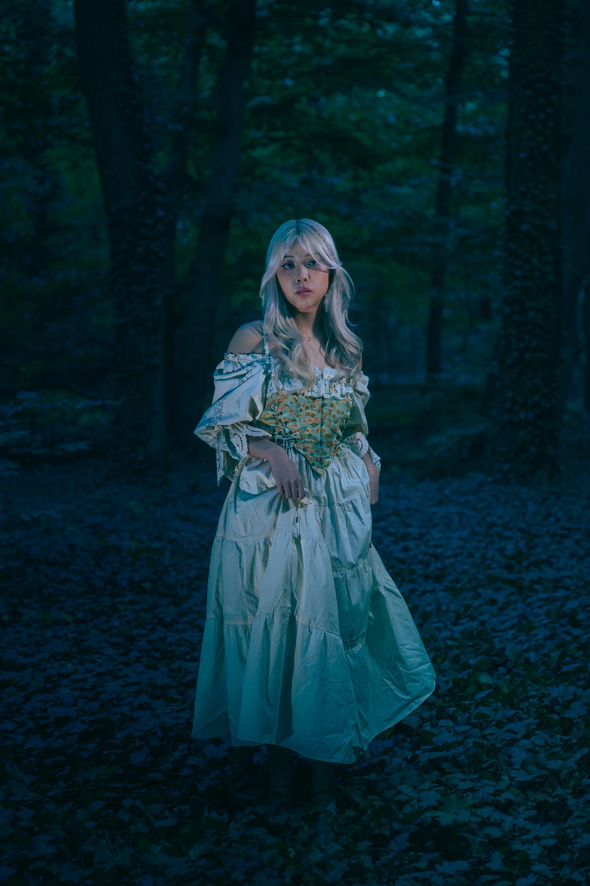 A woman with long blonde hair in a flowy dress standing in a forest at night, with blue lighting creating a mysterious atmosphere. Cinematic portrait photography