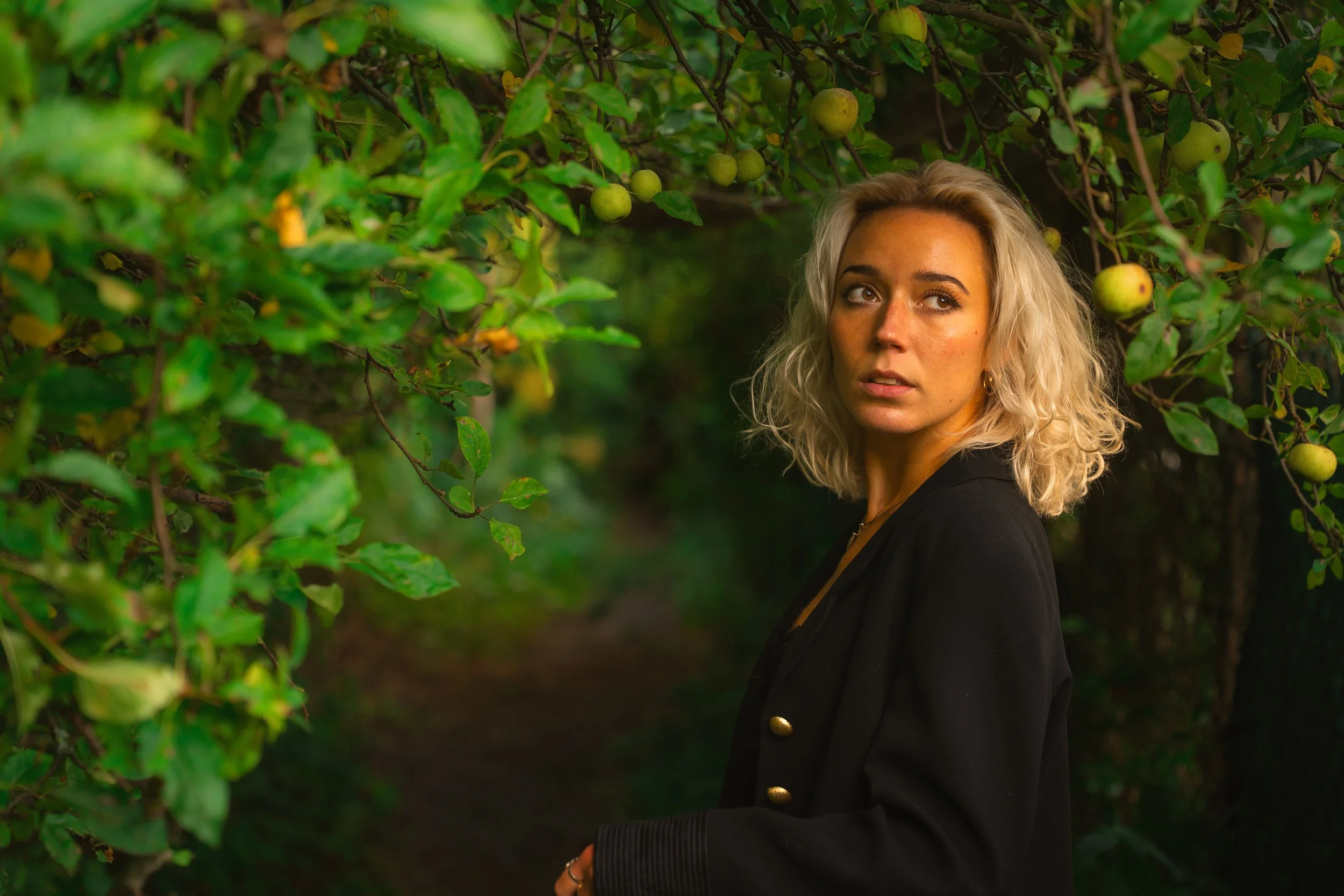 A woman with blonde, curly hair standing in an orchard surrounded by green apple trees. Portrait photography
