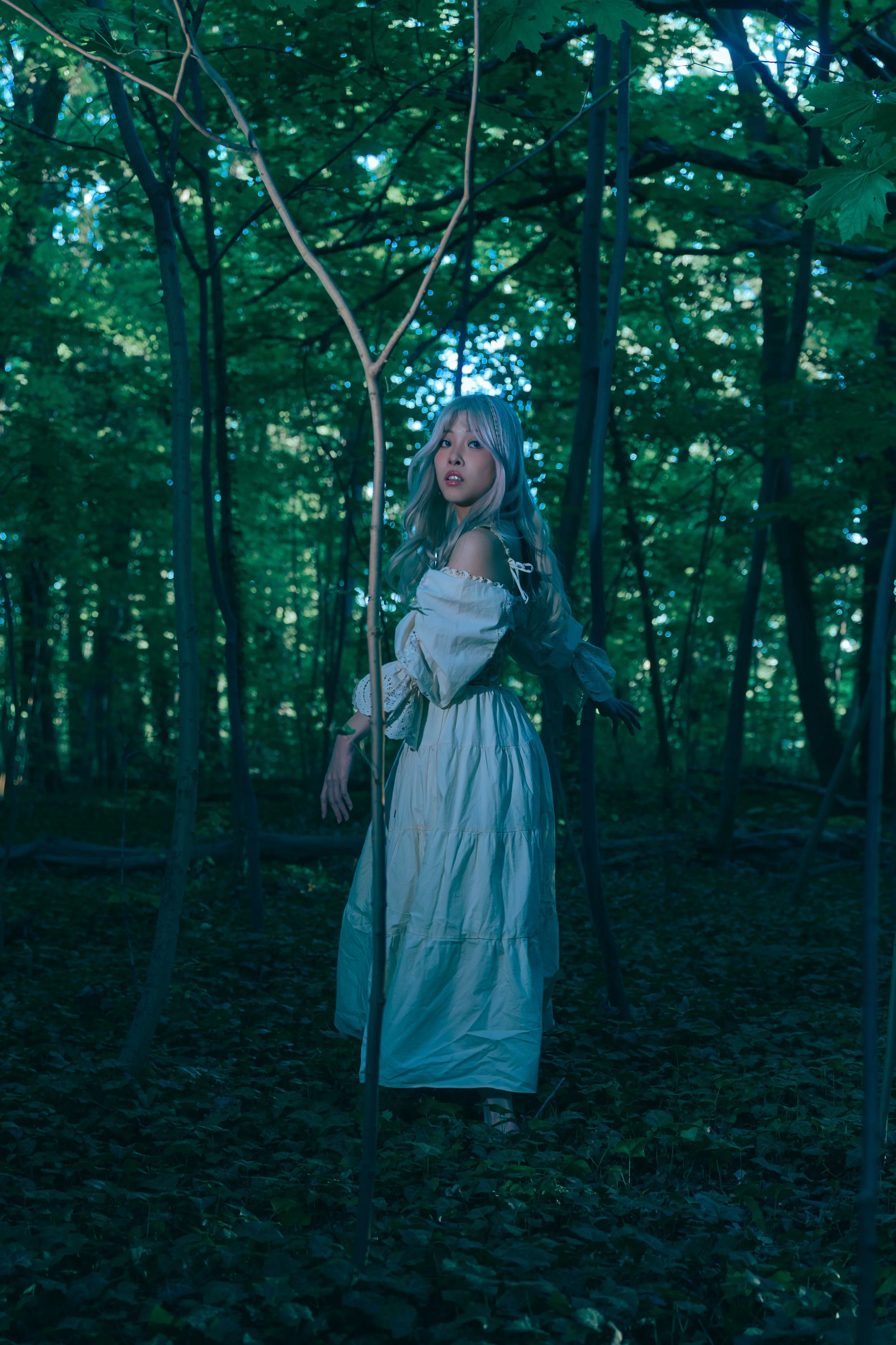 A woman with long, light-colored hair in a white dress stands in a dense, green forest with tall trees surrounding her. Cinematic portrait photography