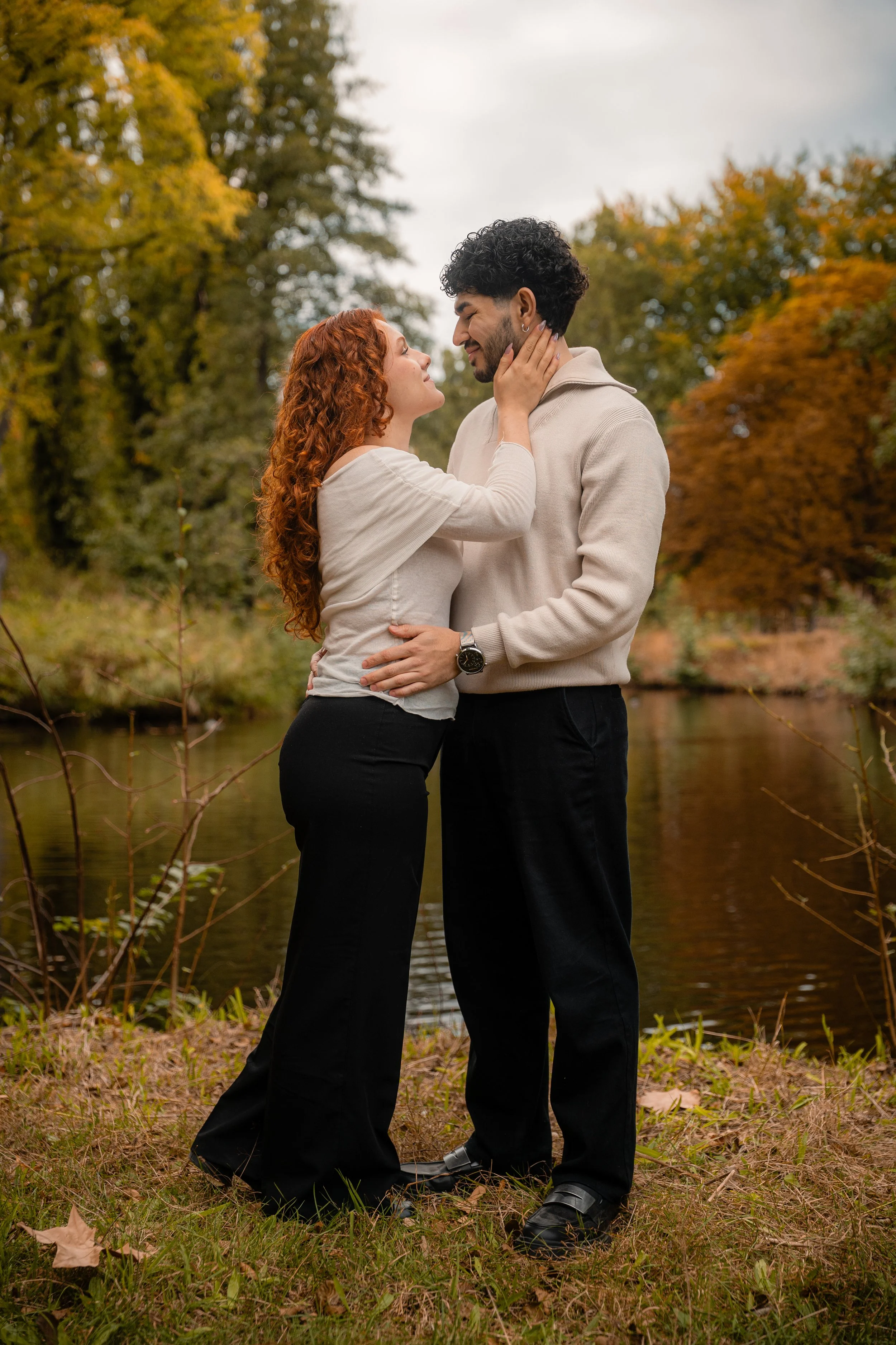 A couple standing close together outdoors by a river, embracing and gazing into each other's eyes, with autumn trees in the background. Couple photography
