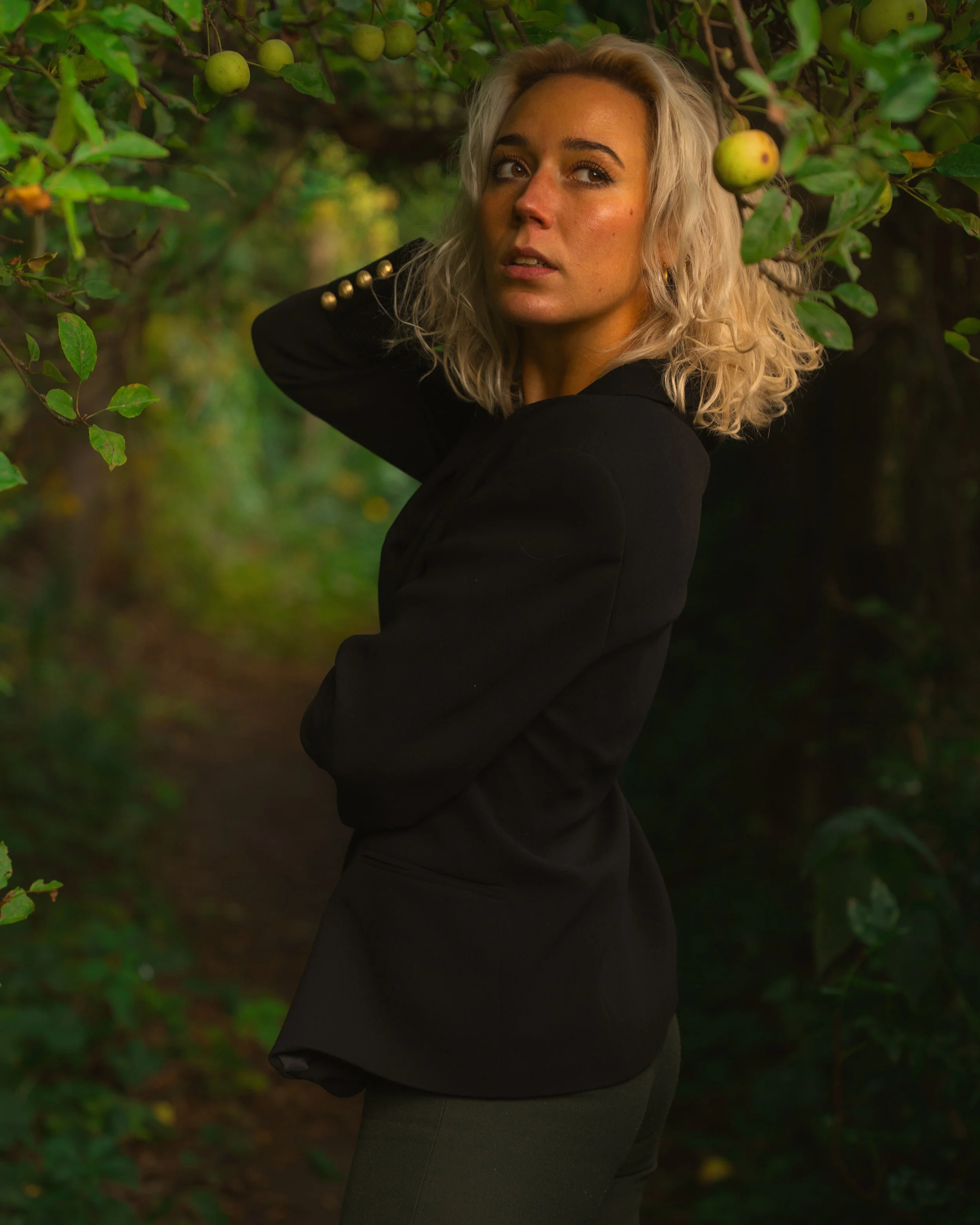 A woman with blonde curly hair wearing a black jacket with gold buttons standing among green apple trees. Portrait photography