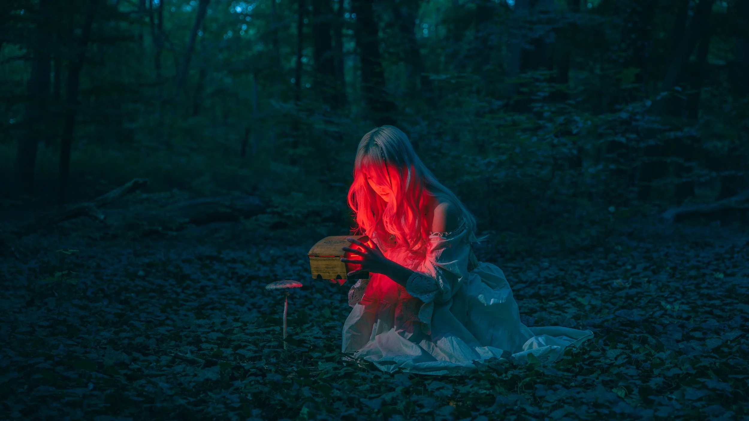 A woman in a white dress kneeling in a dark forest, illuminated by a red glow from a small box she is holding. Cinematic portrait photography