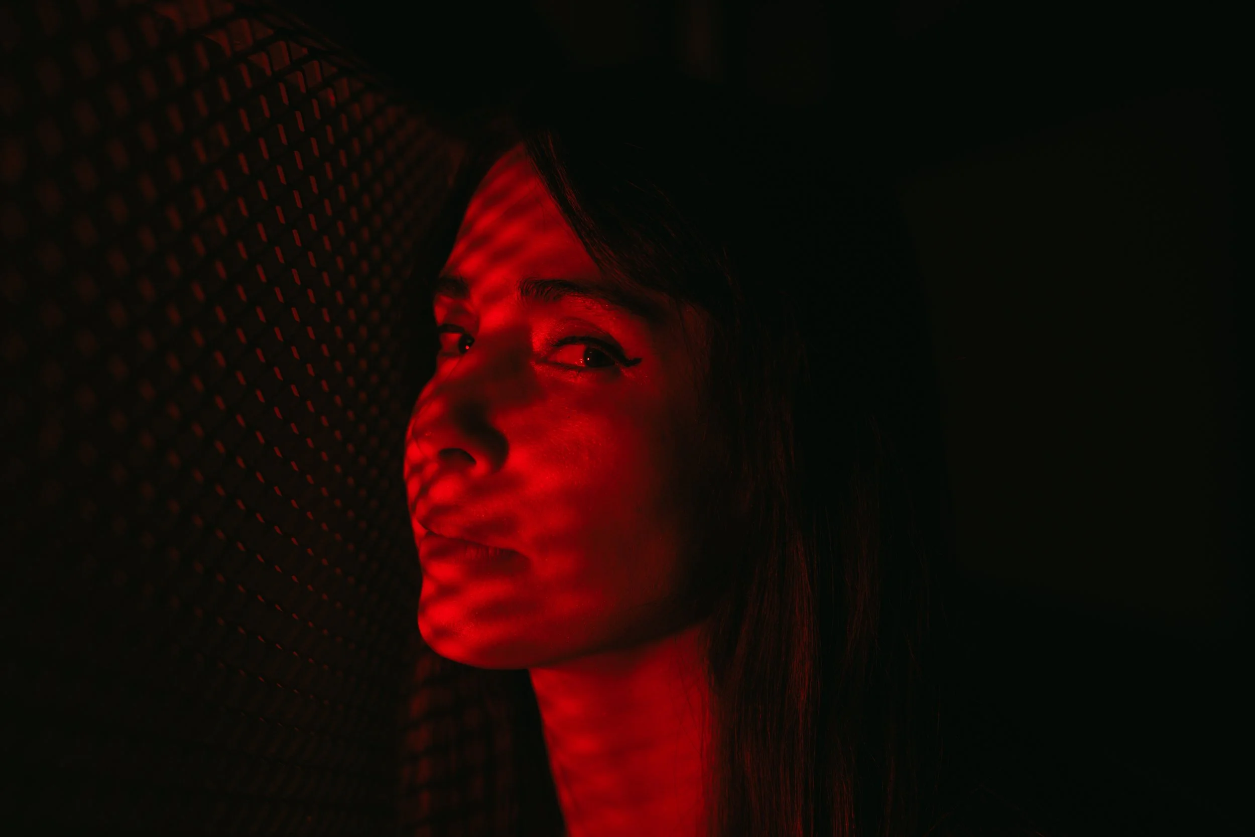 A woman bathed in red light with a patterned shadow across her face, looking sideways in a dark environment. Cinematic portrait photography