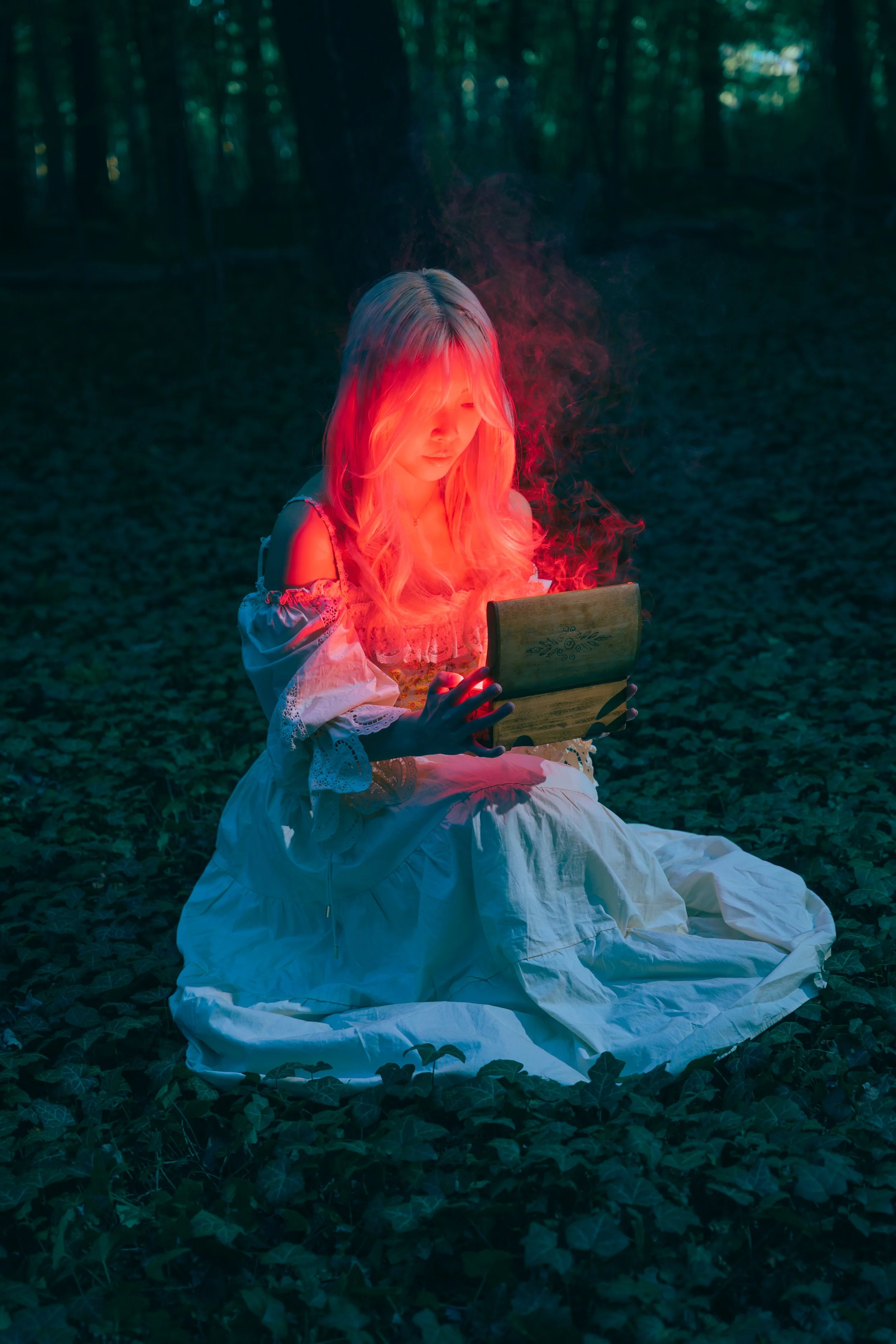 Young woman dressed in a white Victorian-style dress sitting on the ground in a dark forest, holding an open wooden box emitting red and blue glowing smoke. Cinematic portrait photography