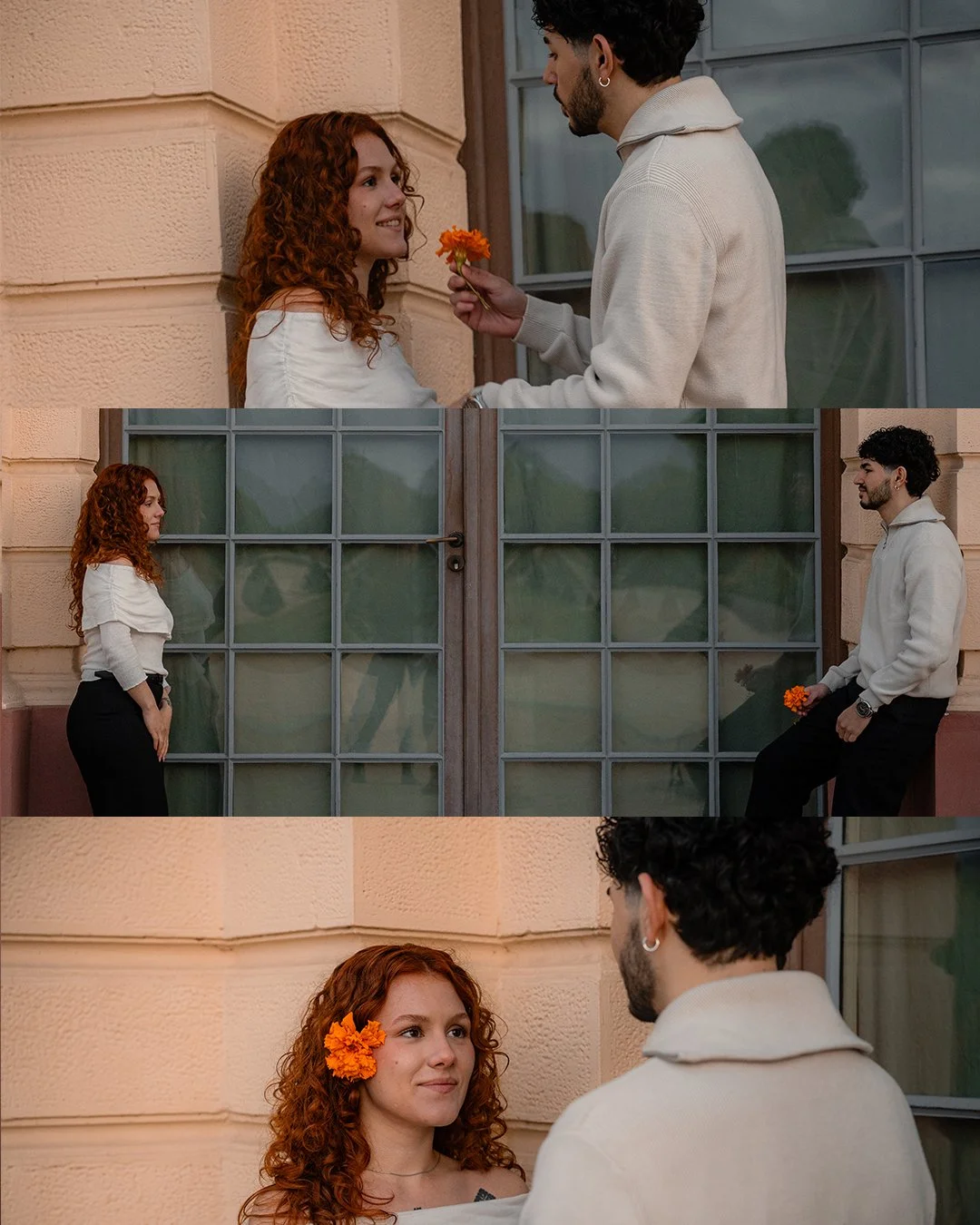 A young woman with curly red hair and a flower in her hair smiling at a young man with dark curly hair, who is holding an orange flower and looking at her. They are standing outside a peach-colored building with large windows, engaging in a romantic