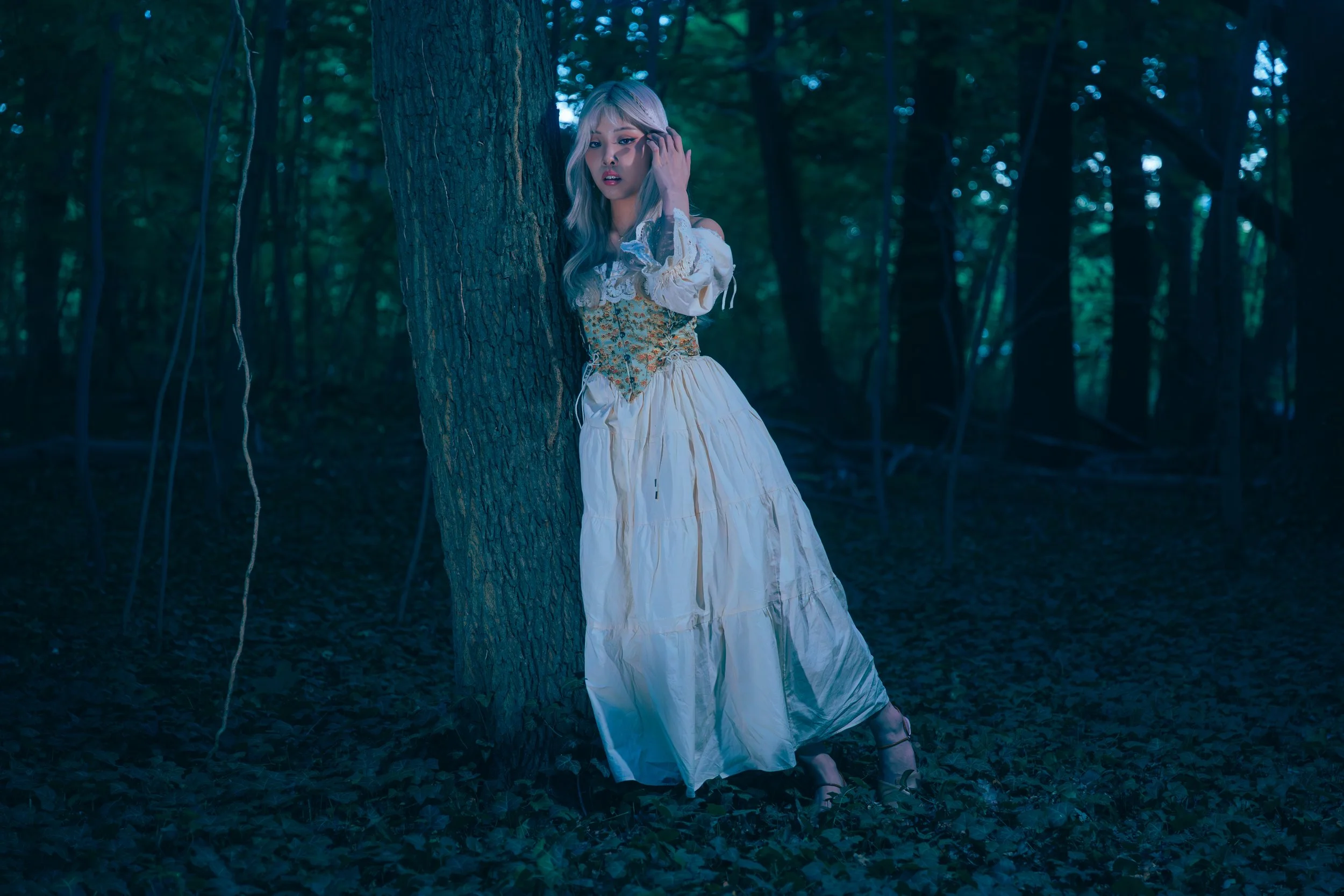 A woman with blond hair wearing a vintage style white dress with floral detailing, standing in a dark forest leaning against a tree, with a surprised or worried expression. Cinematic portrait photography