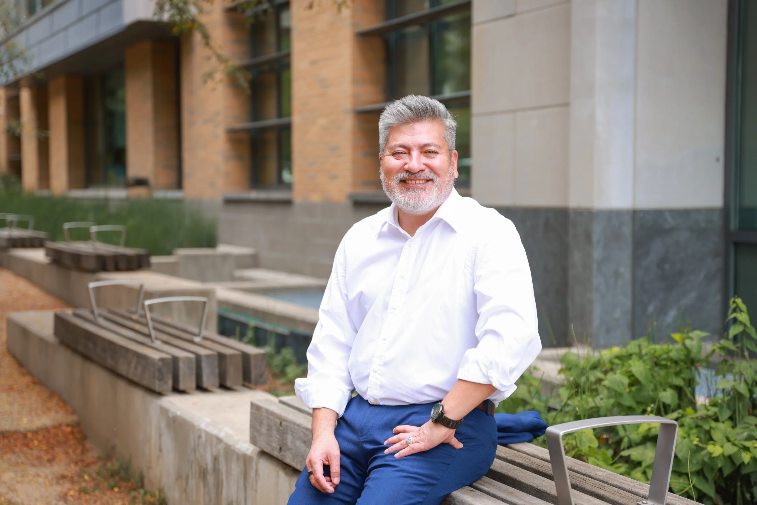 A smiling middle-aged man Juan Sosa with gray hair and a beard, dressed in a white shirt and blue pants, sitting on a bench outdoors in front of a modern brick and concrete building with large windows, greenery surrounding the area.