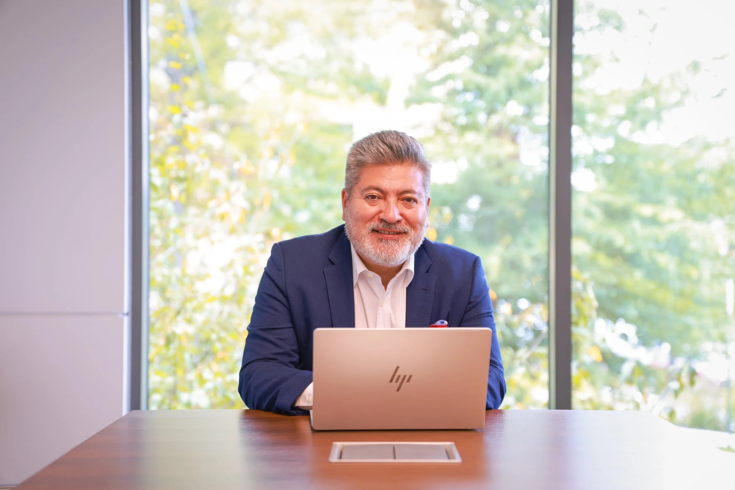 A smiling middle-aged man Juan Sosa with gray hair and beard, wearing a navy blazer and white shirt, sitting at a desk in front of a laptop in a bright office with large windows and green trees outside.