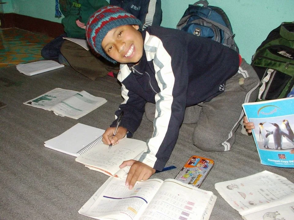 A smiling boy wearing a colorful knit hat and dark jacket is lying on the floor, writing in notebooks. There are textbooks and school supplies around him, including a penguin booklet and a school bag.