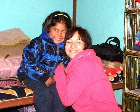 A young girl and a woman smiling and hugging in a room with bed and shelves.