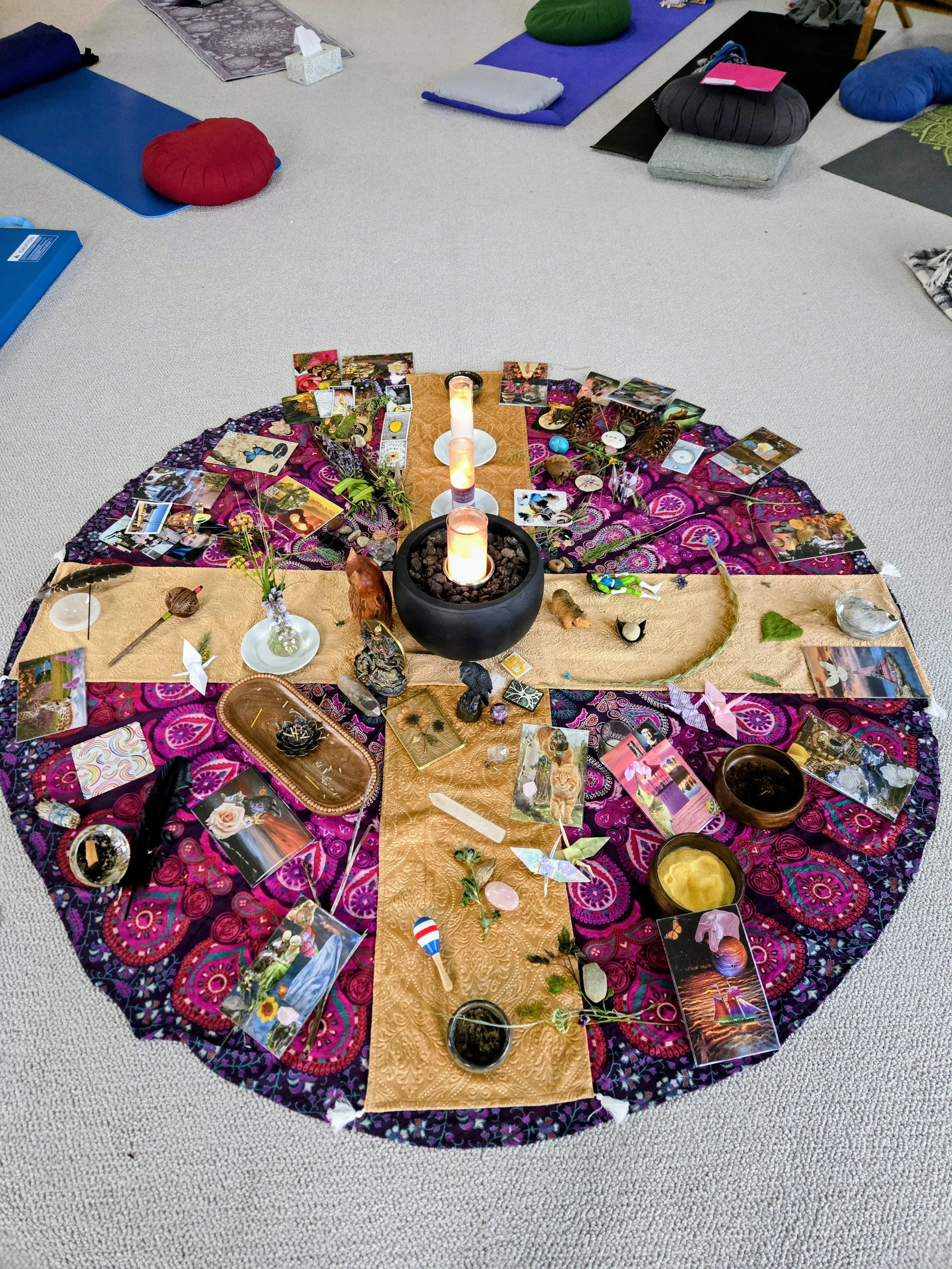 A circular altar with various items and pictures on a patterned cloth in a room with yoga mats and cushions. The center features candles in a black bowl, surrounded by crystals, herbs, and decorative objects.