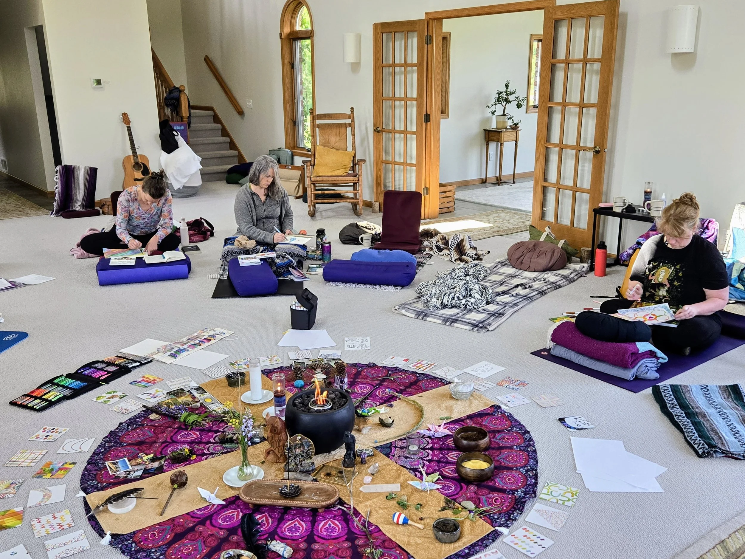 Three women sitting on the floor engaging in an arts and crafts activity with various supplies, blankets, and pillows around them in a spacious room with open French doors and a decorative altar with candles and figurines in the center.