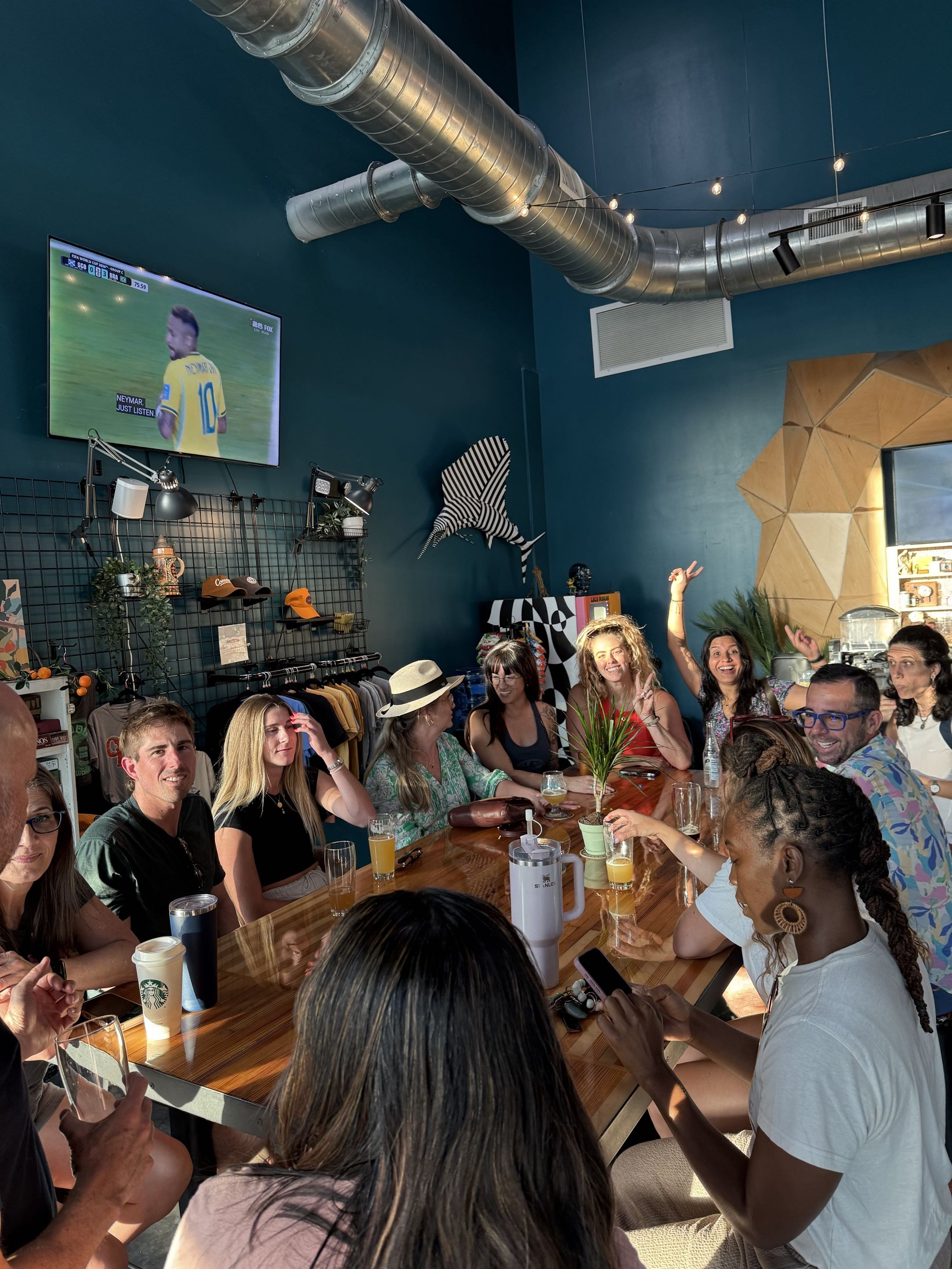 People gathered around a long wooden table in a brightly lit room with dark blue walls. Some individuals are smiling and engaging with their phones, beverages, and conversations. Visible decor includes hats, a striped shirt, plants, and wall-mounted artwork. A large TV screen shows a soccer match.