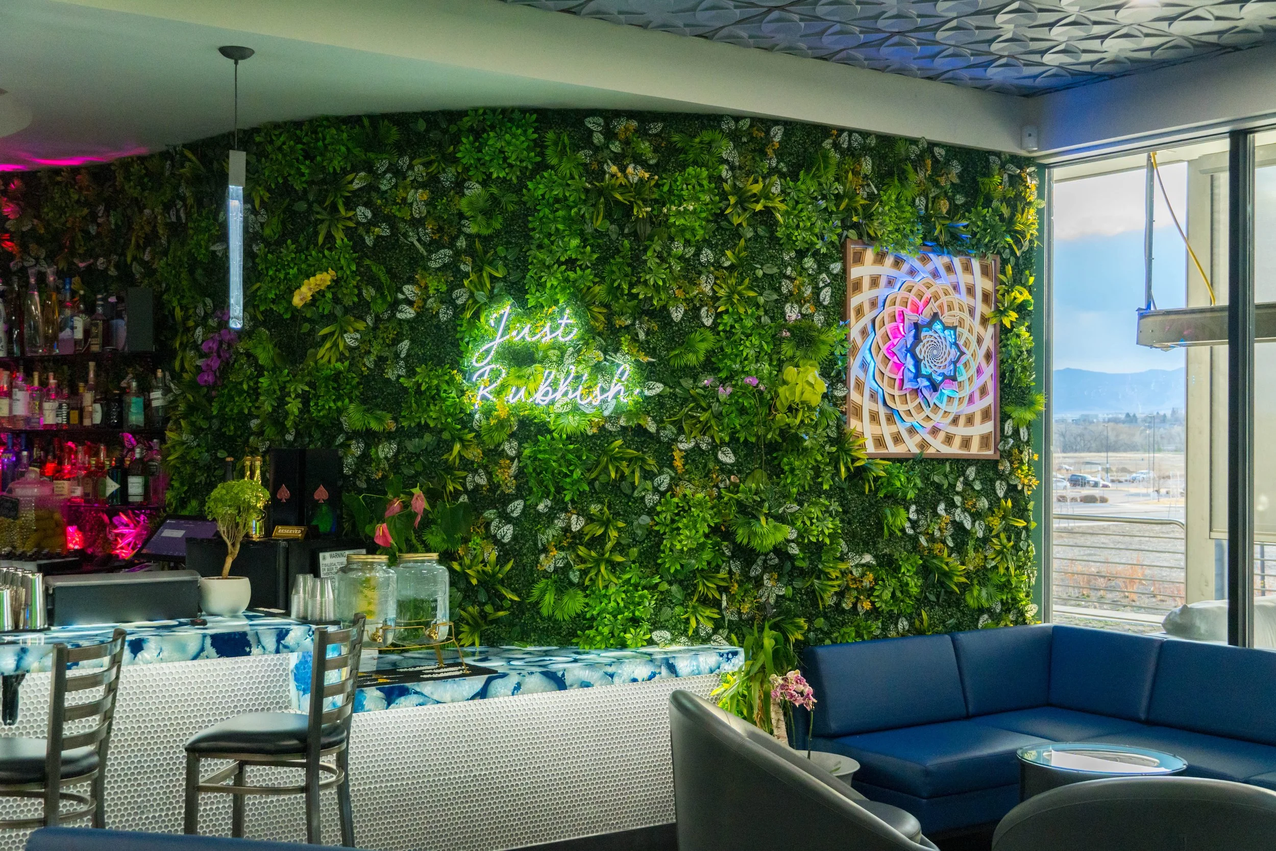 Interior of a lounge with a green living wall decorated with a neon sign that says 'Just Rubbin,' a blue sofa, a table, and a large window showing an outdoor parking lot and mountains.