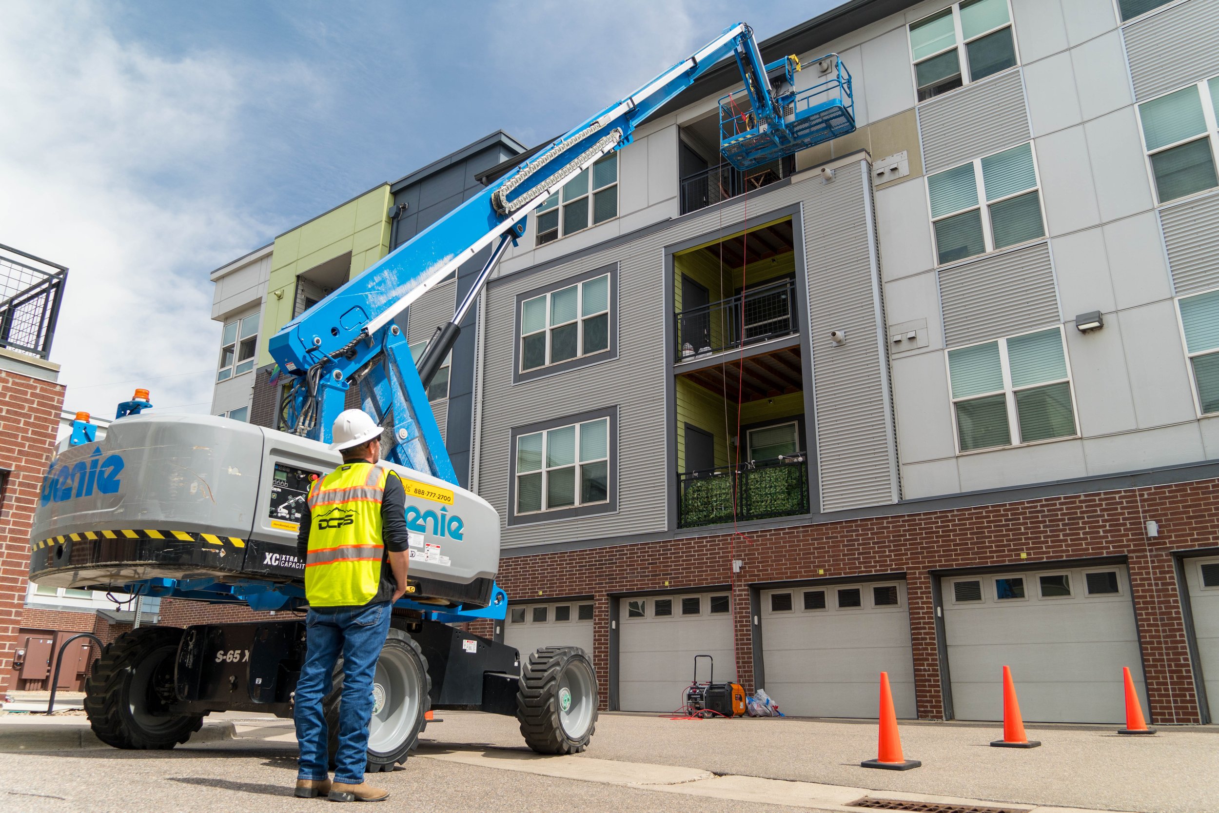 A construction worker in a white helmet and yellow safety vest stands next to a blue mobile crane lifting equipment to the upper floors of a multi-story residential building. The building has a brick lower level and gray and white siding above, with 