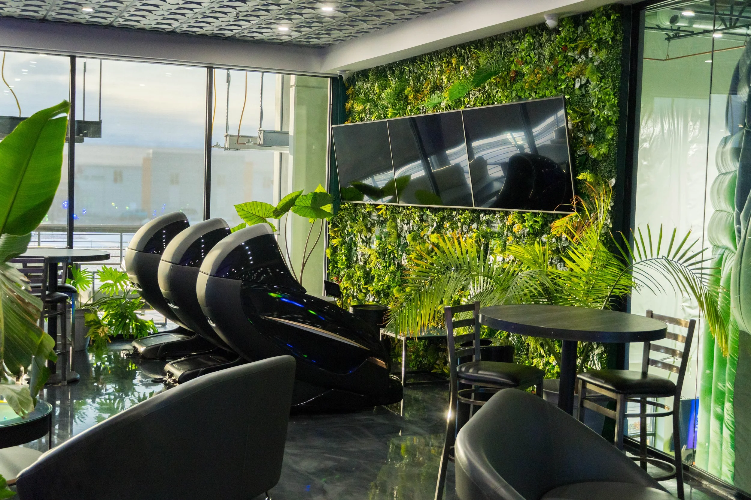Modern indoor space with three black futuristic chairs, a small round table, and lush green plants on a wall and in pots, with large windows letting in natural light.
