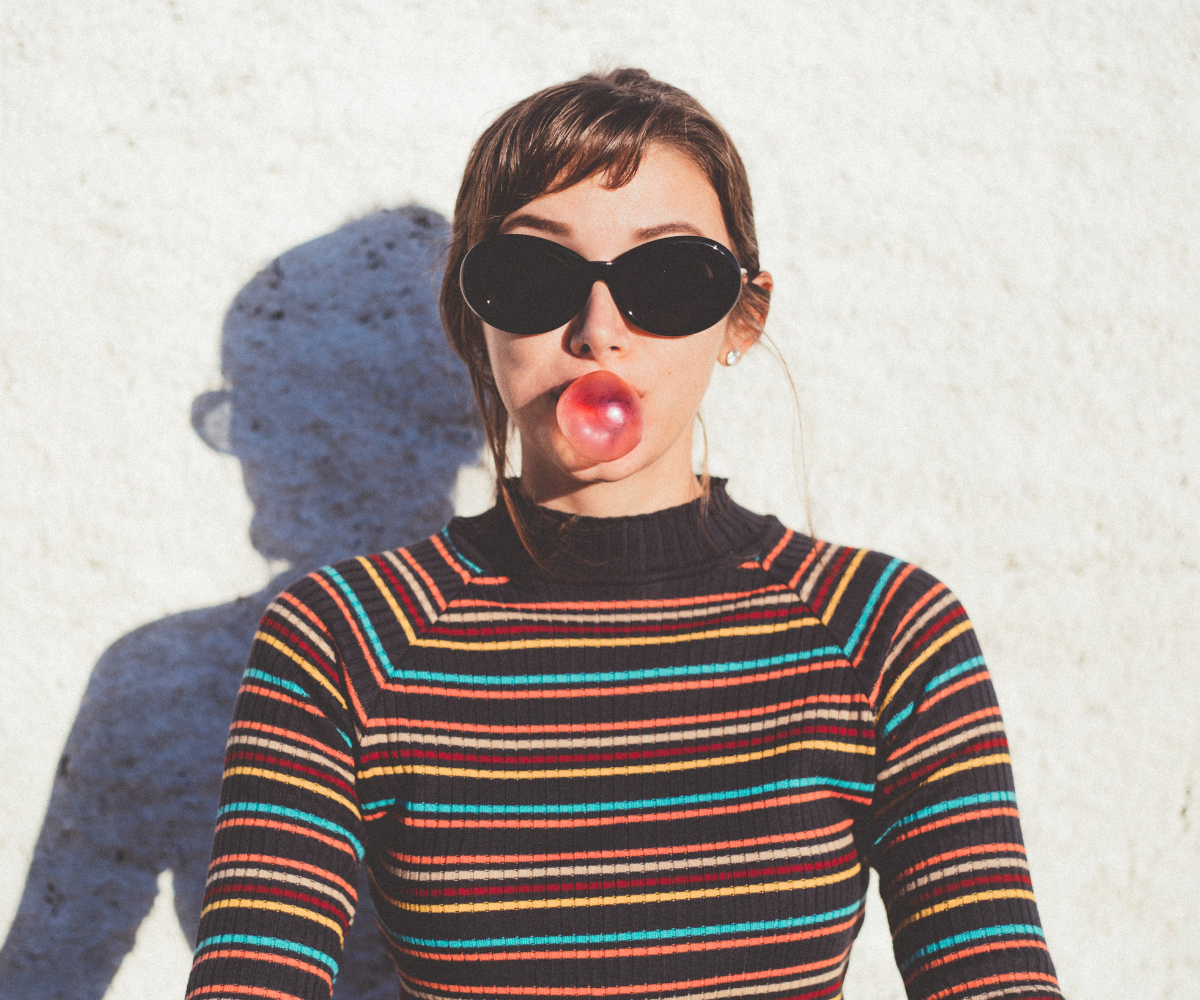 Naine, kellel on päikeseprillid ja värviline triibuline kampsun, puhub nätsust mulli, seistes valge seina ees ja heites varju.