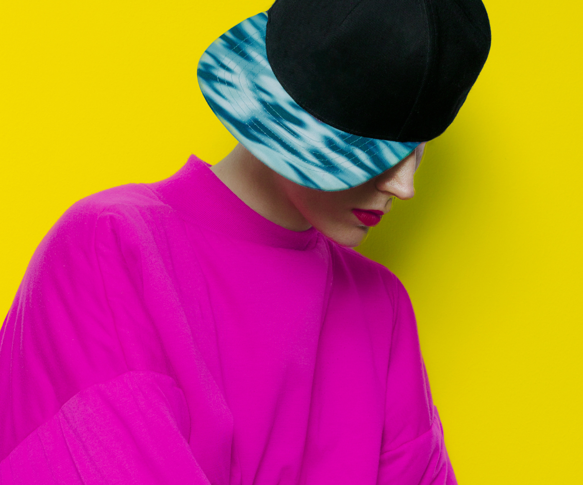 Naine, kellel on seljas erksaroosa topp ja peas must müts sinise-valge mustrilise nokaga, seisab kollase tausta ees.