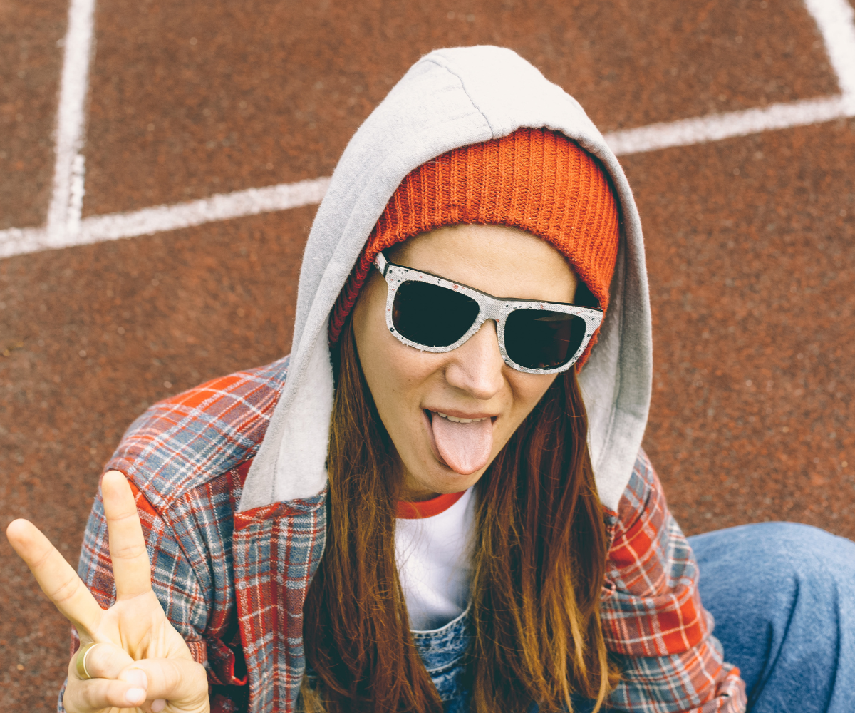 Naine, kellel on päikeseprillid, punane müts ja dressipluus, teeb rahumärki ja ajab keelt välja, olles õues punasel jooksurajal.