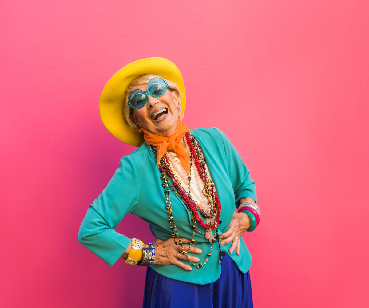 Eakas naine, kellel on peas suur kollane müts, seljas türkiissinine jakk ja mitu värvilist kaelakeed, naerab käed puusal, roosa tausta ees.