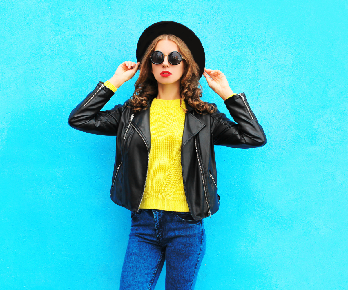 Noor lokkis juustega naine seisab erksinise seina taustal, kandes musti päikeseprille, laia servaga musta kübarat, musta nahktagi, kollast kampsunit ja siniseid teksaseid.