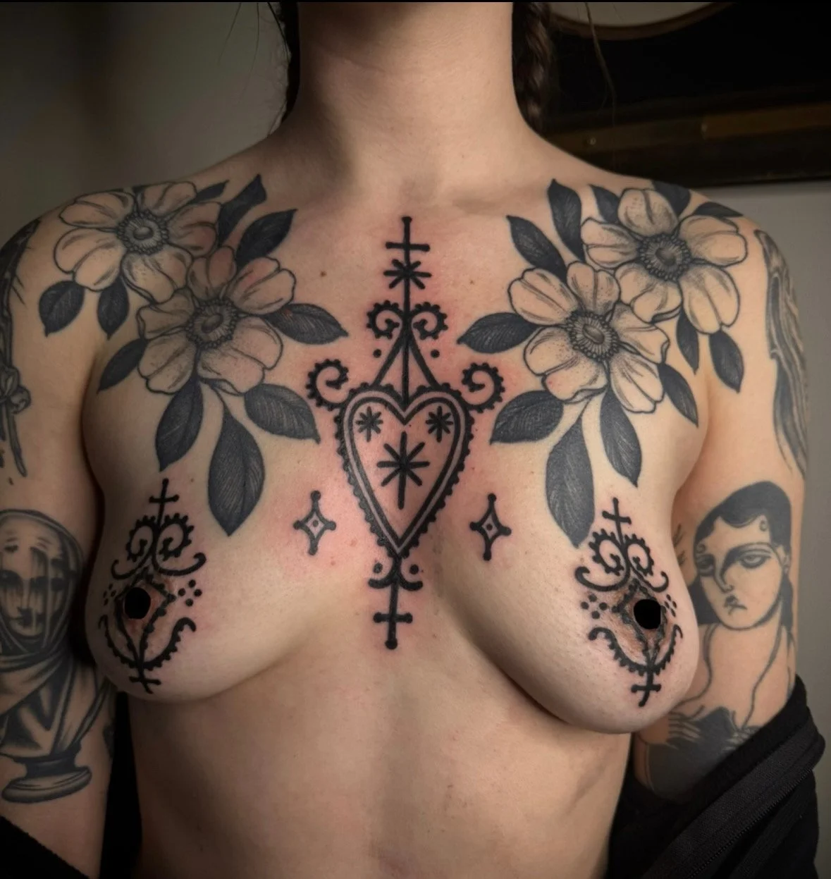 Close-up of a woman's chest with black tattoos, including flowers on the shoulders, controlled symbols, and a large heart with intricate designs and lines.