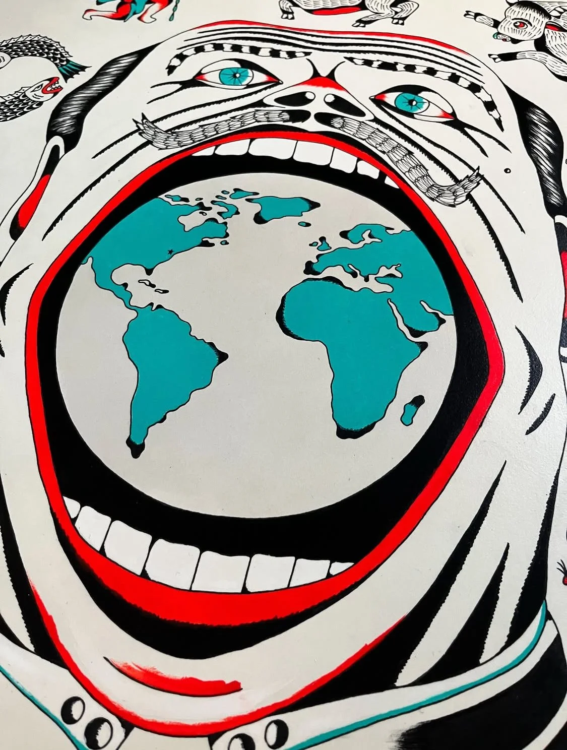 Artwork featuring a large face with piercing blue eyes, open mouth revealing white teeth, with the globe inside the mouth. The face has black and white features with red accents, and surrounding elements include a snake and other decorative motifs.