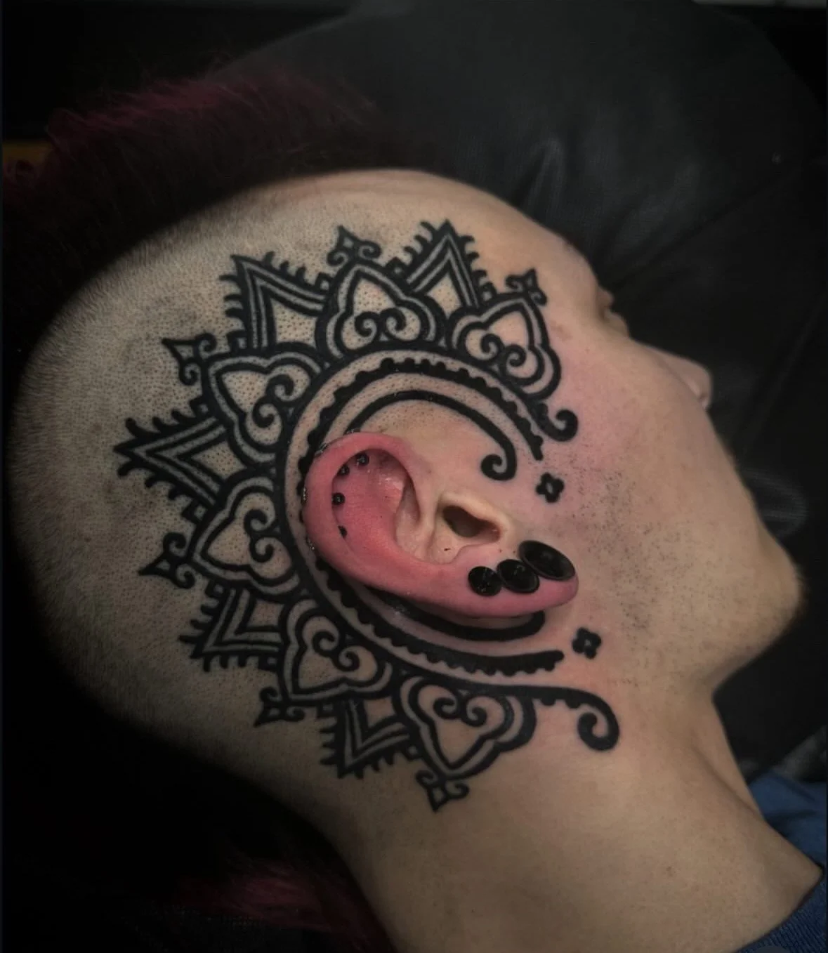 A person's head with a large decorative black tattoo behind the ear, and multiple piercing with black jewelry in the ear.