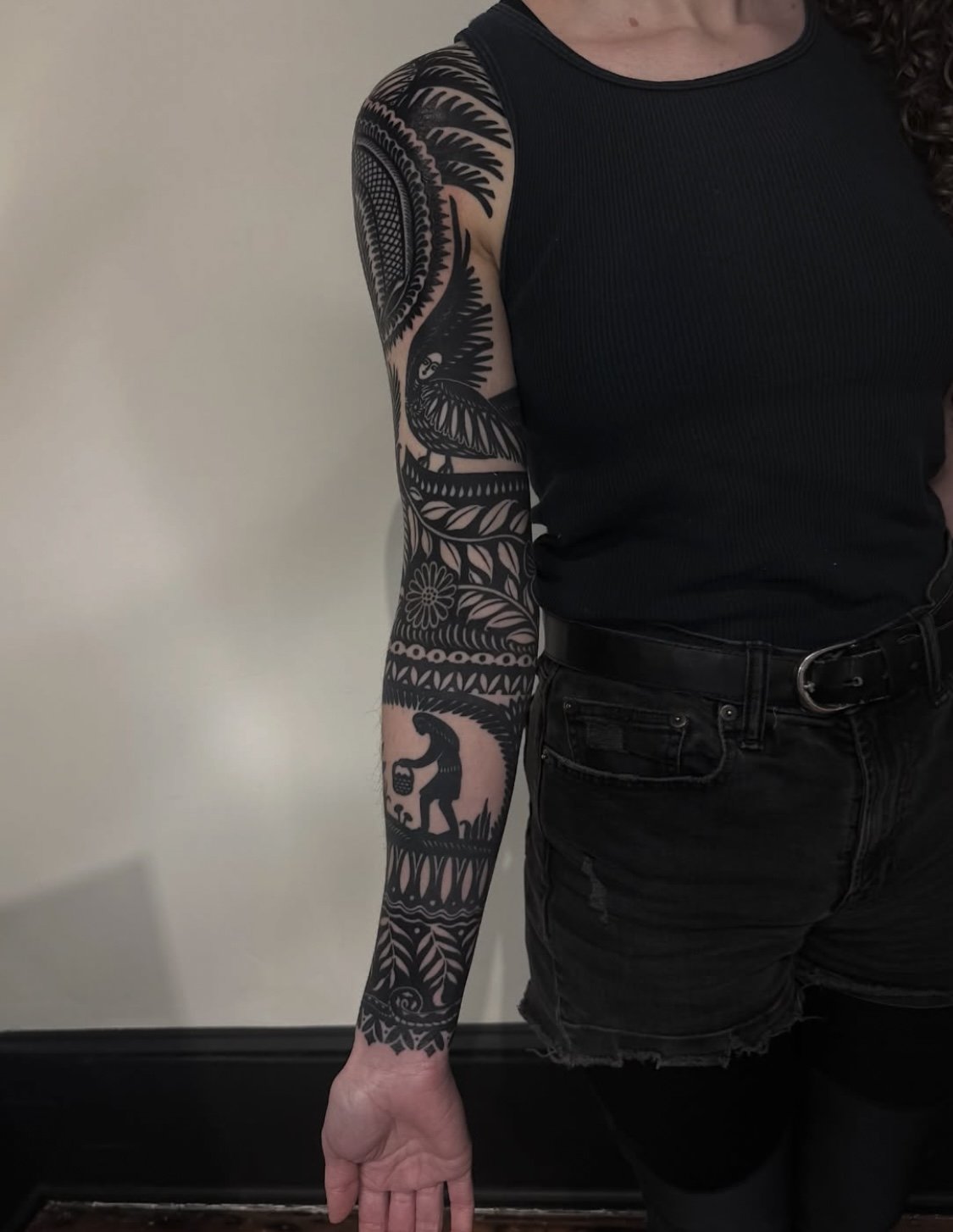 A person with a black and gray tattoo sleeve on their right arm, featuring a palm tree, bird, and smaller designs, standing against a beige wall. They are wearing a black sleeveless top and black jeans with a belt.