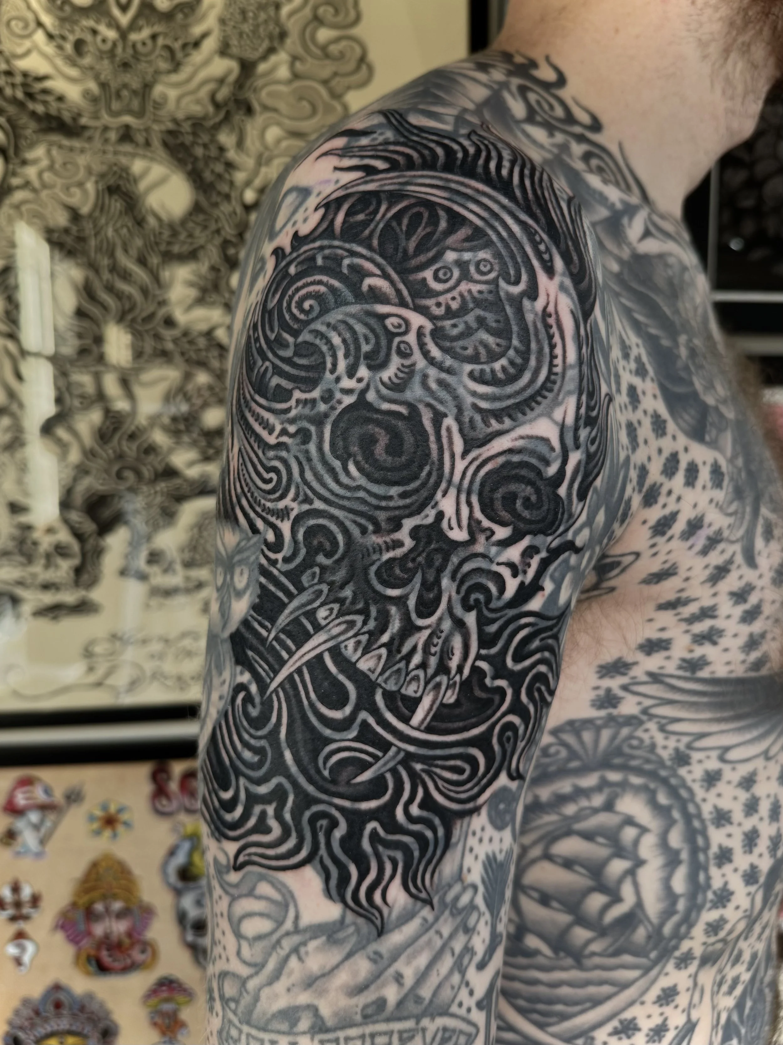 Close-up of a person's shoulder and upper arm with detailed black and gray tattoo artwork of a skull with intricate patterns and designs.