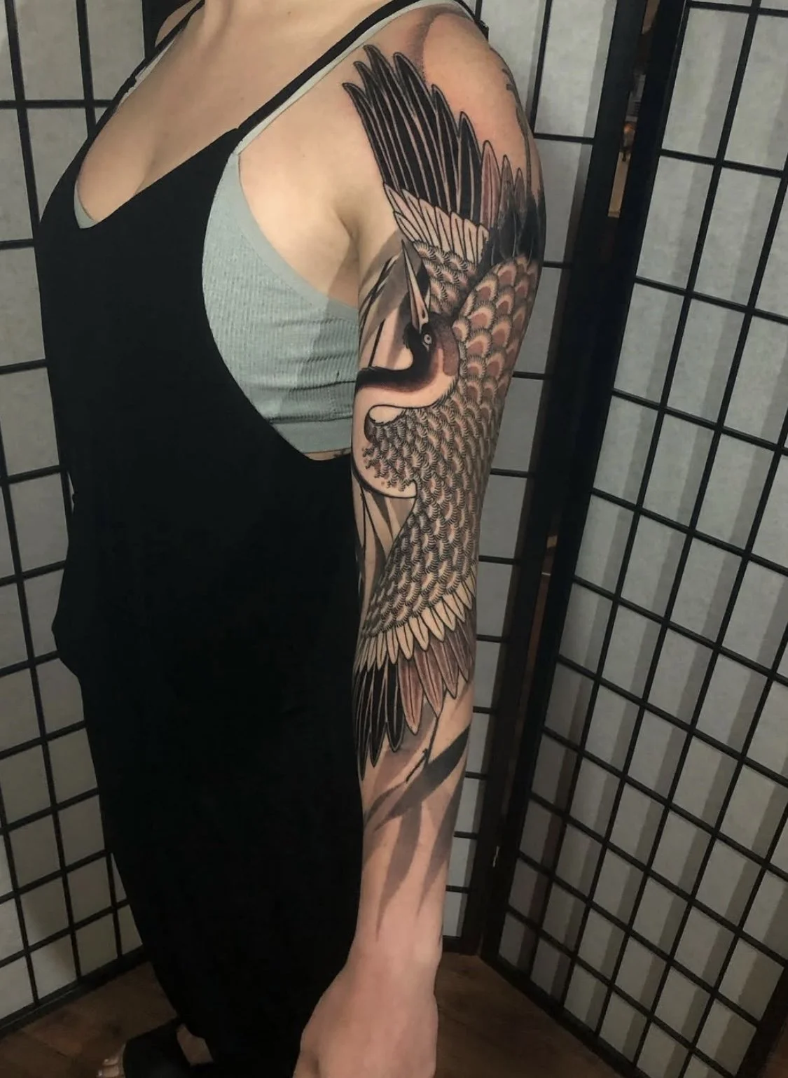 A person with a large black and gray tattoo of a bird on their upper arm and shoulder, standing in front of a folding screen.