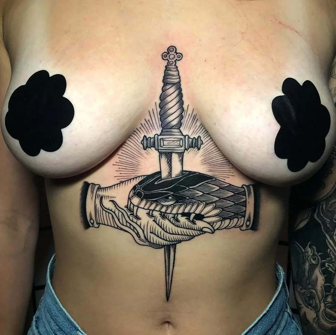 Tattoo of a sword piercing through a hand holding a fish, with black cloud-like shapes covering the nipples, on a person's chest.