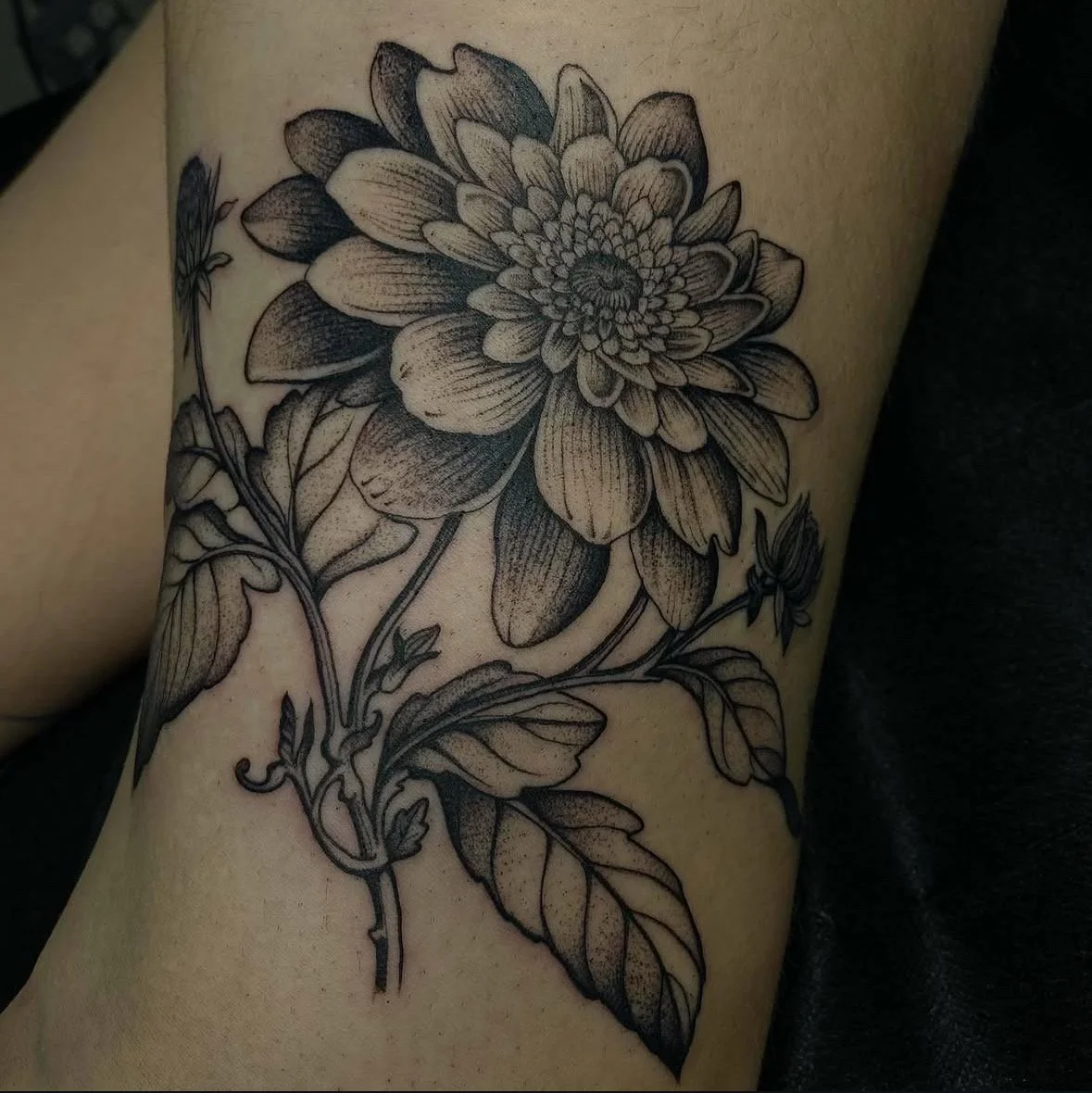 A detailed black and grey tattoo of a large flower with many petals, accompanied by a few leaves and smaller buds.