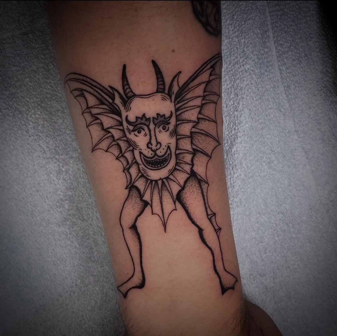 Tattoo of a demon-like face with horns, whiskers, large ears, and a wide smile, surrounded by spider web design elements, on a person's arm.