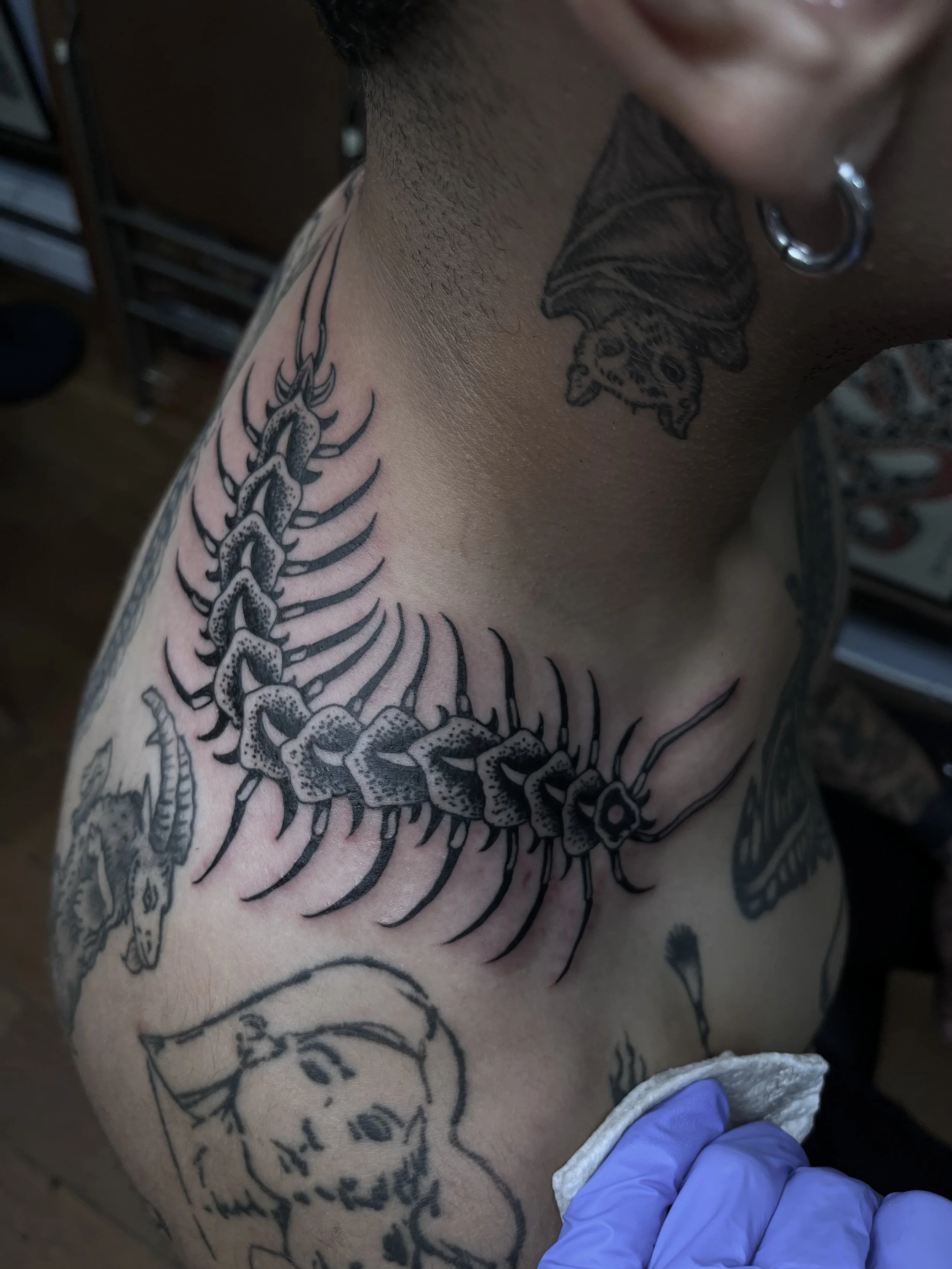 A person's shoulder and upper chest with black ink tattoos, including a centipede tattoo on the shoulder and other designs, with a person wearing a purple glove cleaning or preparing the skin for tattooing.