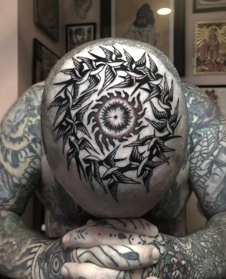 Tattooed person with a bald head, showcasing a black and gray tattoo of birds and a circular design in the center, surrounded by framed artwork in the background.