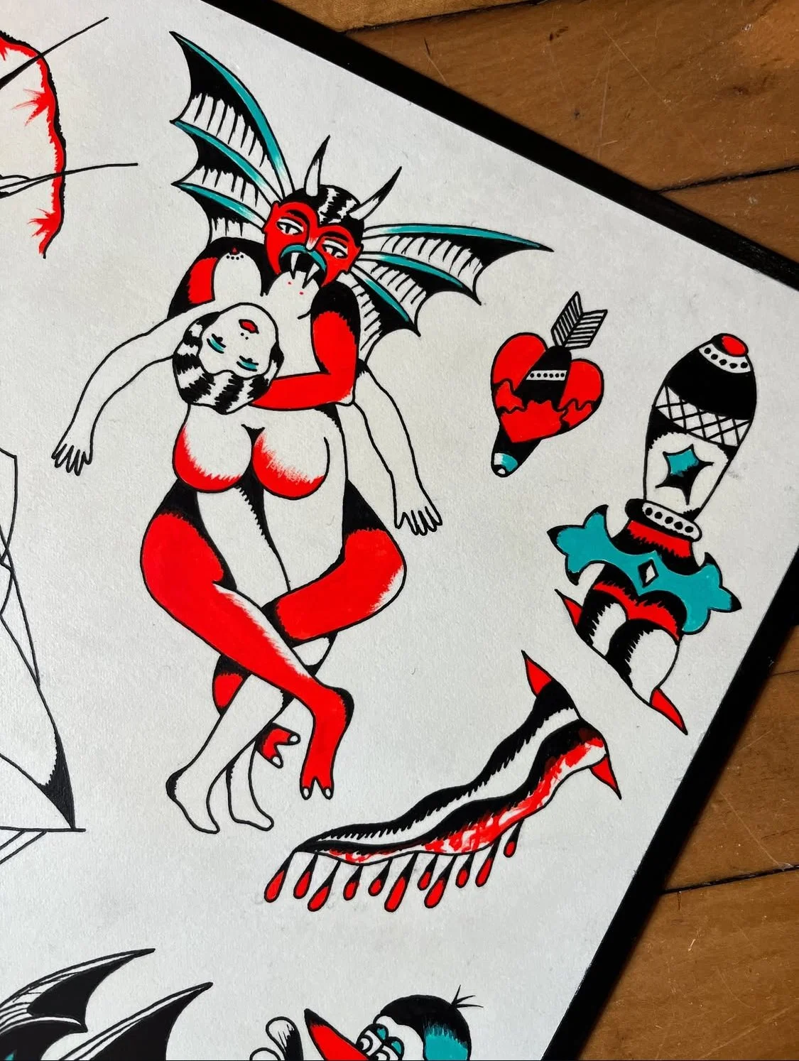 Tattoo-style artwork depicting a mythological creature with a red and black face, bat-like wings, and a human female with closed eyes, lying in the creature's arms. Surrounding elements include a heart with an arrow, a decorated spear, and abstract p