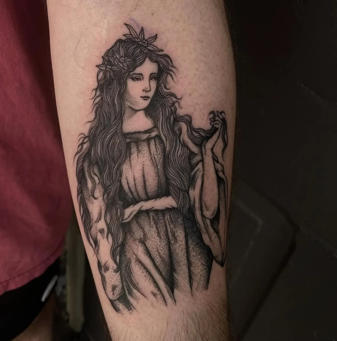 Black and gray tattoo of a woman with long, wavy hair holding a branch, wearing a crown of leaves, with detailed shading and line work.
