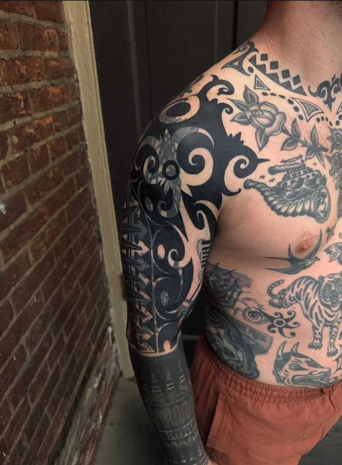 Close-up of a person's upper body with extensive black and gray tattoos on arm, chest, and shoulder, featuring swirling patterns, floral motifs, a tank, an eye, a tiger, and various other designs. The person is wearing orange shorts, standing against